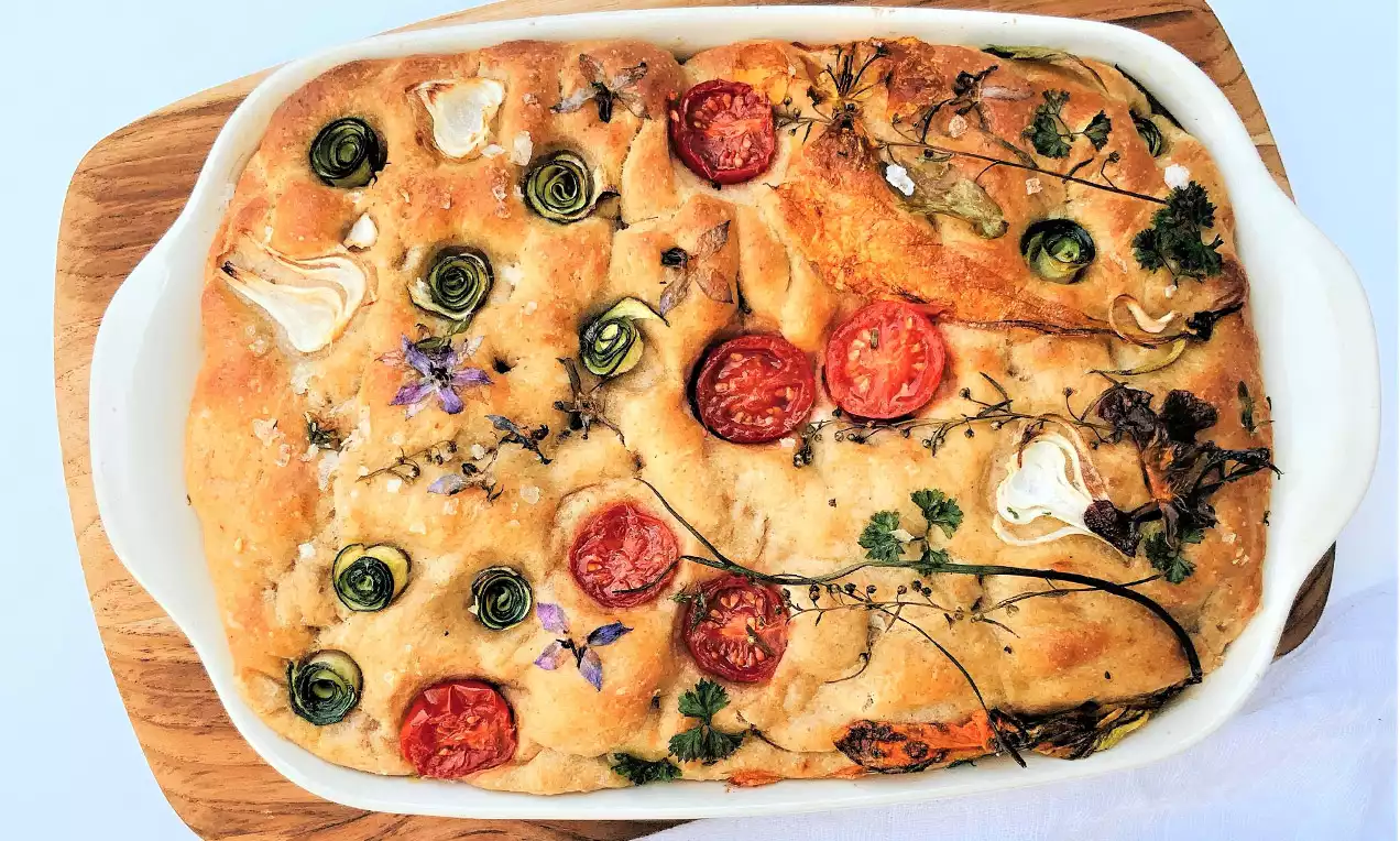 Zlatnožuta focaccia s hrskavom koricom, ukrašena svježim rajčicama, srebrnasto-zelenim tikvicama i grančicama začinskog bilja na bijelom pekaču