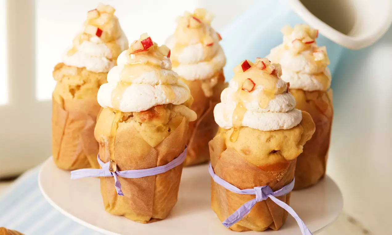 Cupcakesi svijetlozlatne boje s kremom od šlaga, komadićima pečenih jabuka i kapima meda, ukrašeni ljubičastim trakama na stalku