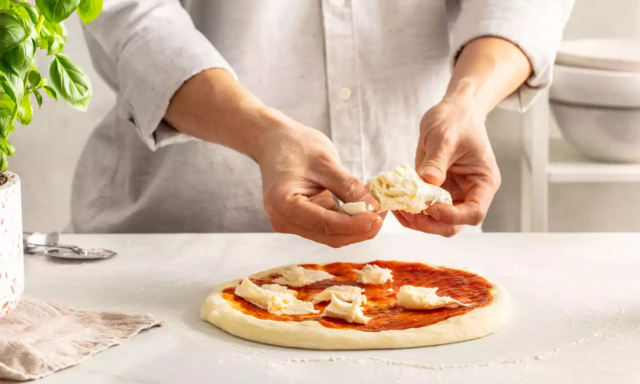 Pizza od kiselog tijesta s tankom, zlatnom korom prekrivena crvenim umakom od rajčice, komadićima svijetlog sira i svježim bosiljkom na bijelom stolu