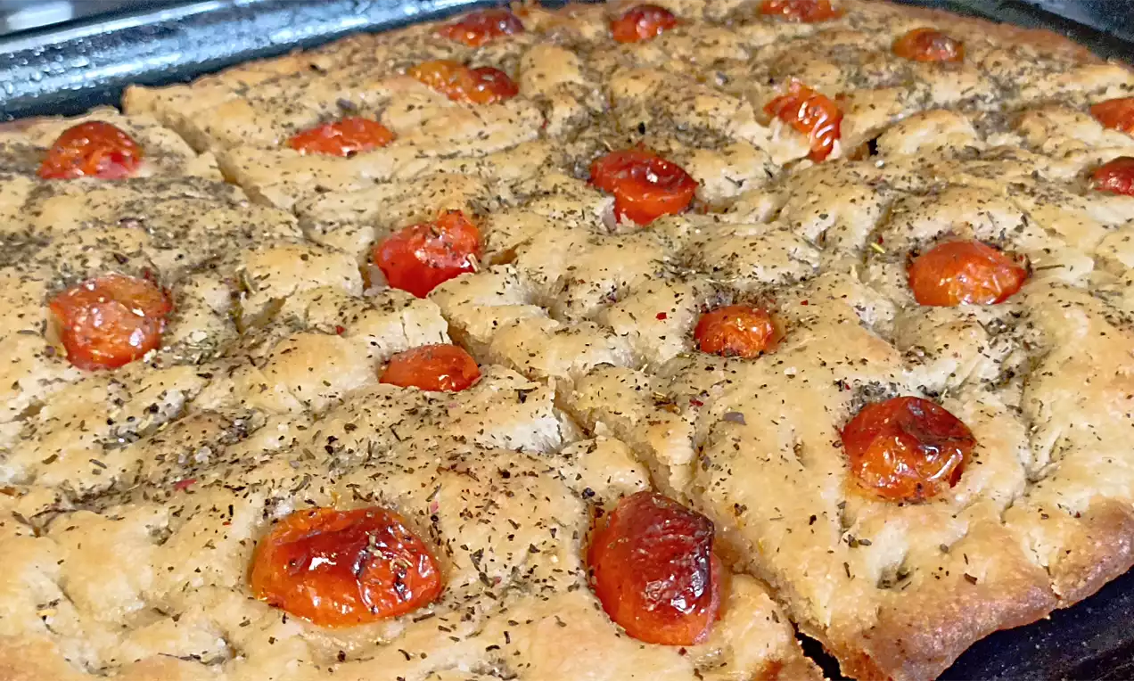 Zlatnosmeđa focaccia s hrskavom korom, prekrivena pečenim crvenim cherry rajčicama i začinskim biljem na tamnom plehu