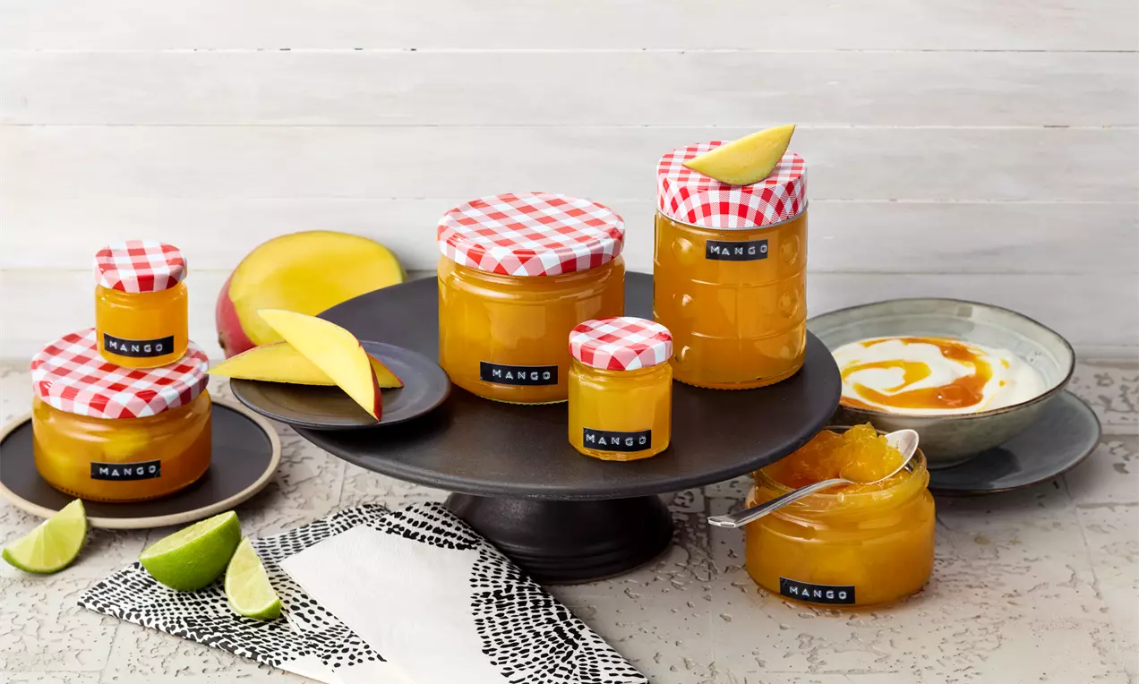 Staklenke zlatnožute marmelade s crveno-bijelim poklopcima na tamnom postolju, uz kriške manga, limete i tanjur s marmeladom