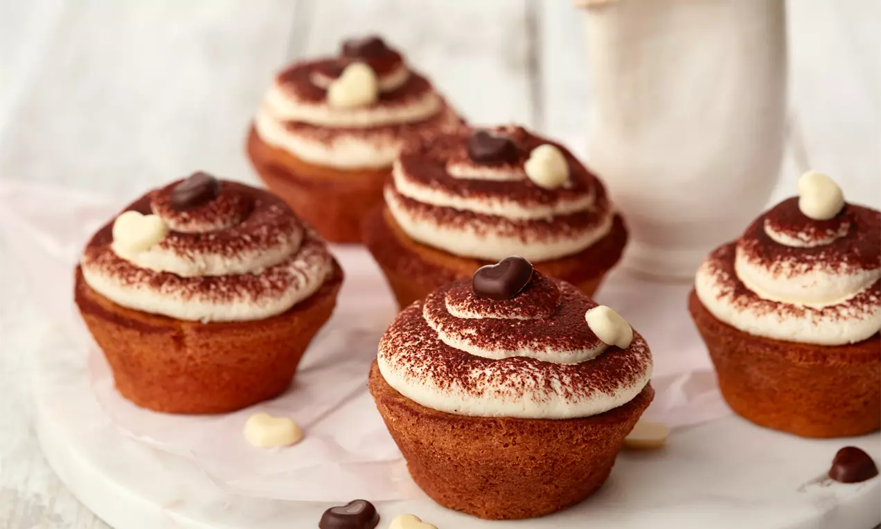 Smeđi cupcakes s kremastim bijelim vrhom posutim kakaom, ukrašeni čokoladnim srcima na bijelom tanjuru uz maleni vrč