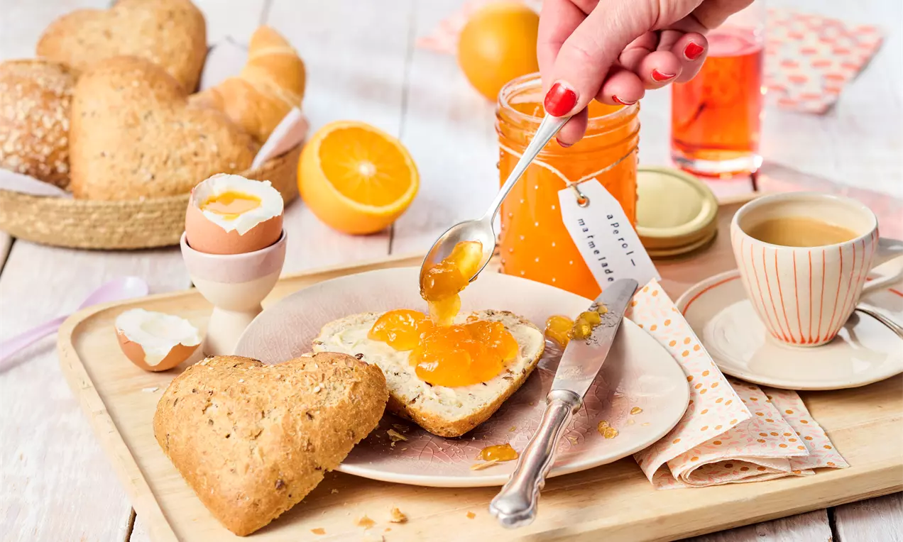 Svijetlonarančasta marmelada s glatkom teksturom razmazuje se na prerezano srcoliko pecivo, okruženo svježim narančama i doručkom