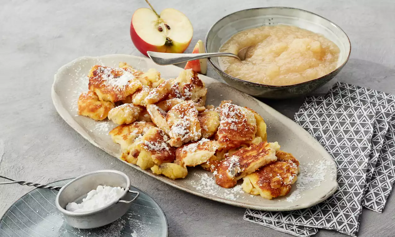 Zlatnosmeđi, nepravilni komadi Kaiserschmarrna posuti šećerom u prahu na ovalnom tanjuru uz zdjelicu svijetložutog umaka od jabuka