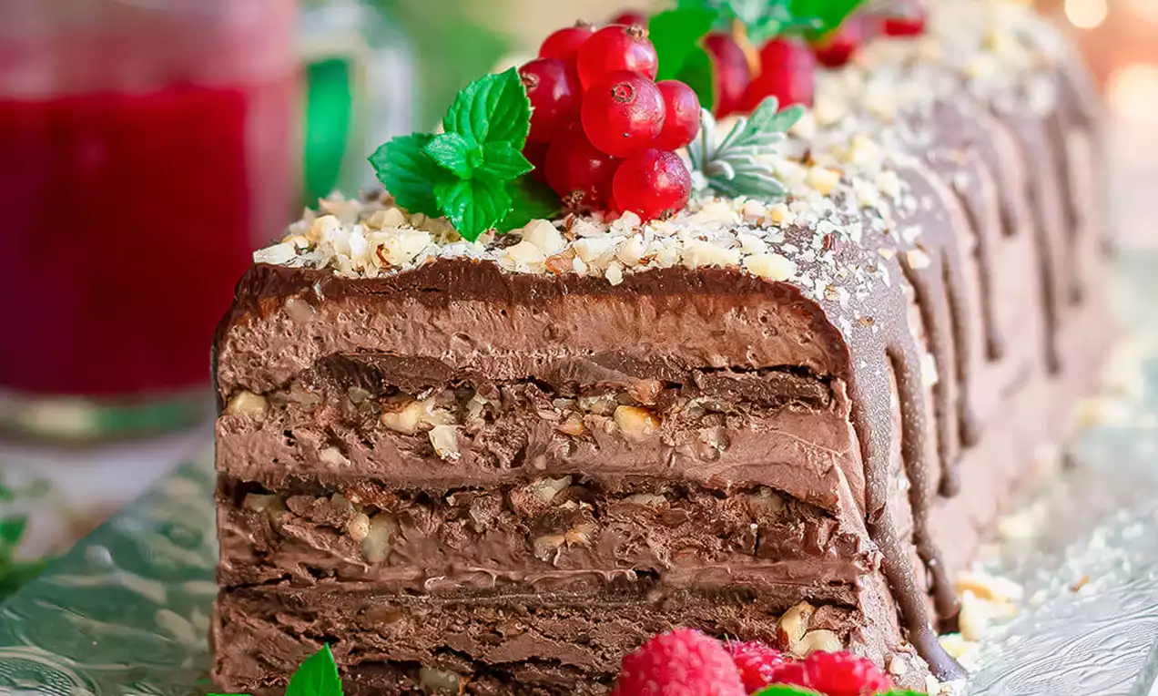 Čokoladni desert pravokutnog oblika sa slojevima kreme i komadićima lješnjaka, ukrašen crvenim voćem, listićima mente i čokoladnim preljevom