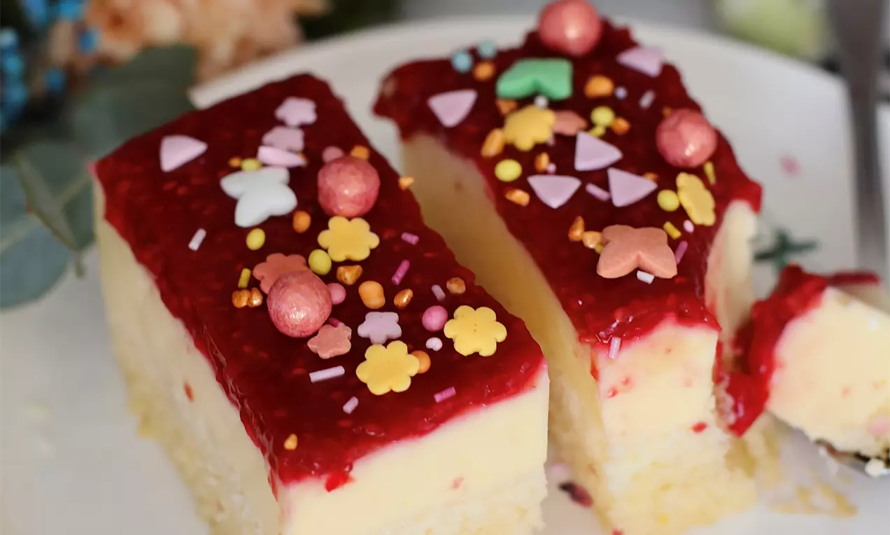 Maline kocke na bijelom tanjuru s žutom kremom na svijetlom biskvitu, prelivene crvenim slojem, posute šarenim dekorativnim mrvicama