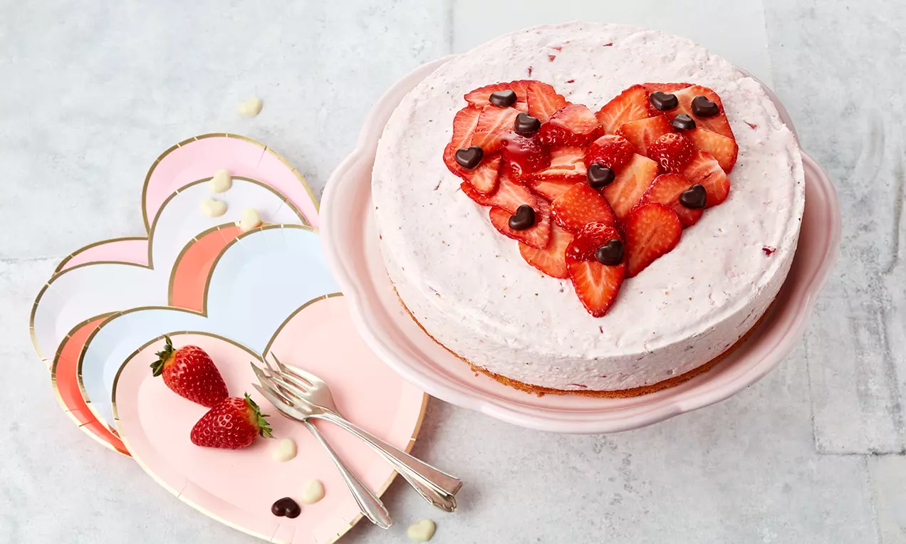Okrugla torta svijetloružičaste kreme na smeđoj podlozi s dekoracijom od svježih jagoda i čokoladnih kapljica u obliku srca na pastelnom stalku