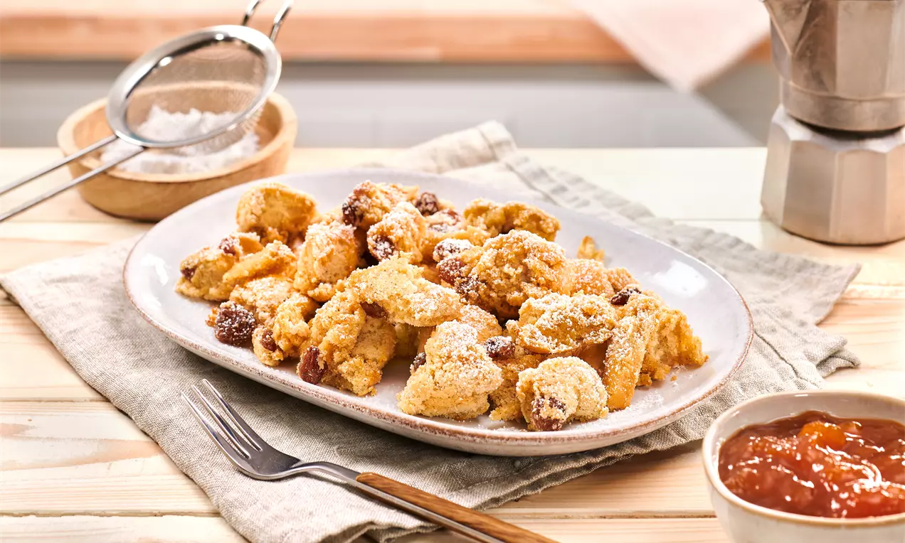 Komadići zlatnosmeđeg Kaiserschmarrna posuti šećerom u prahu, raspoređeni na ovalnom tanjuru uz zdjelicu džema i pribor za jelo