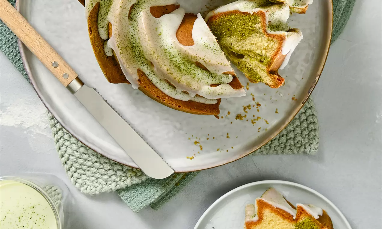 Bundt torta svijetlozelene matcha boje i zlaćanih rubova prekrivena bijelom glazurom, poslužena na bijelom tanjuru uz dva napitka i kriške na manjem tanjuriću