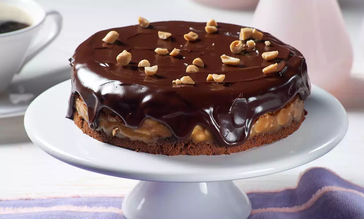 Mala okrugla veganska torta na bijelom stalku s tamnom čokoladnom glazurom, slojem karamele i posutom sjeckanim lješnjacima